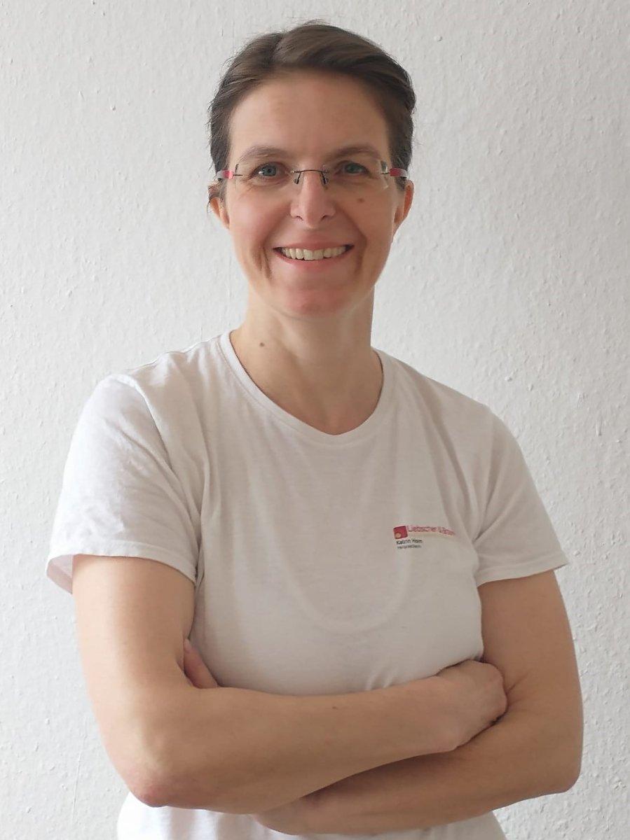 Das Foto zeigt Katrin Horn aus Fürstenwalde / Spree. Sie steht mit einer Schulter an einer weißen Wand gelehnt. Sie hat kinnlange braune Haare und trägt eine rahmenlose Brille. Auf dem Foto trägt sie ein schwarze Kette mit einem dunklen Anhänger, einen sc