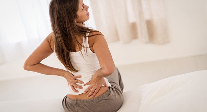 Eine dunkelhaarige Frau, die auf einem Bett sitzt, hält sich mit beiden Armen den unteren Rücken vor Schmerzen.