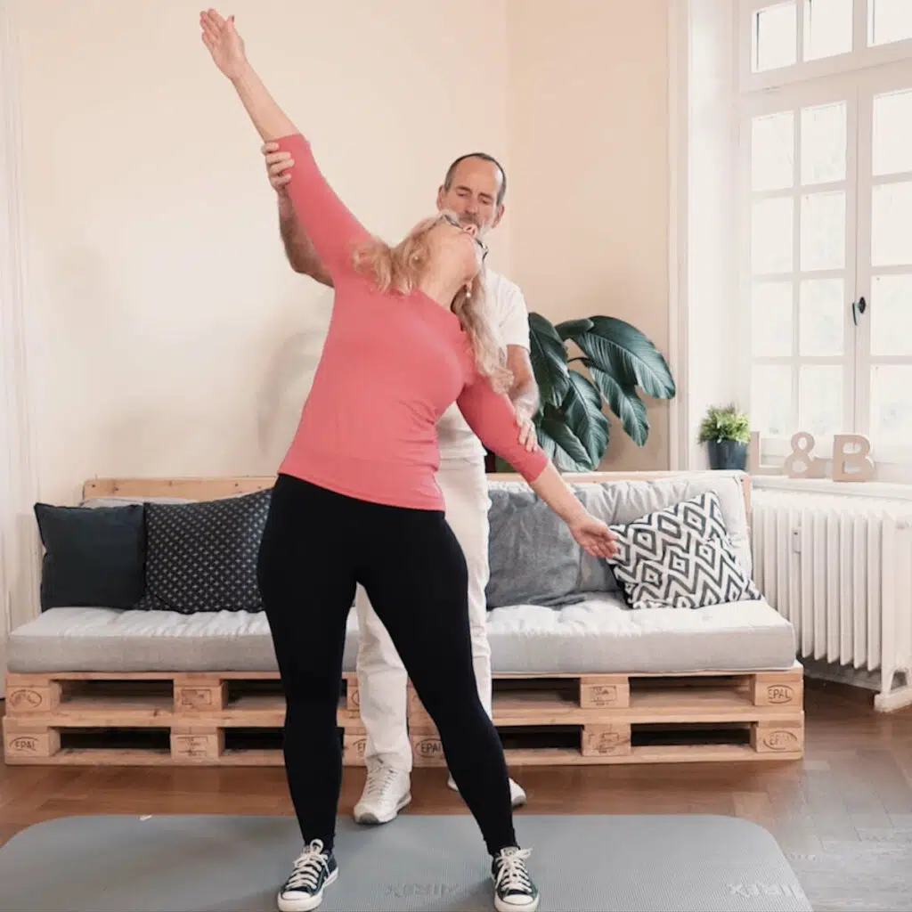 Roland Liebscher-Bracht zeigt einer Frau eine Übung. Sie steht schulterbreit, einen Arm weit über den Kopf gestreckt, den anderen nach unten, sodass beide Arme eine Linie ergeben.