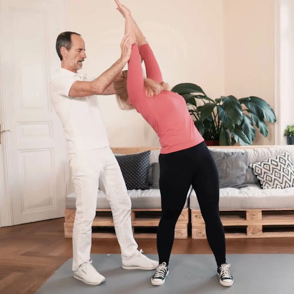 Eine Frau steht hüftbreit auf einer Übungsmatte und macht eine Rotationsübung, indem sie sich mit beiden Armen nach oben gestreckt nach links dreht.