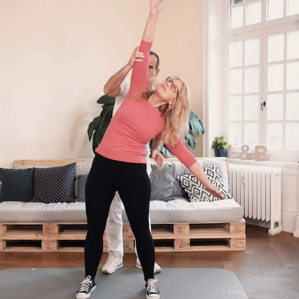 Eine Frau steht hüftbreit auf einer Übungsmatte und streckt dabei den rechten Arm gerade nach oben in die Luft, der linke Arm ist leicht zur Seite gestreckt, um die Übung zu balancieren.