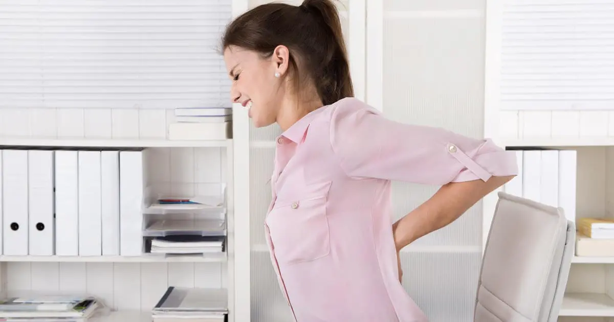 Eine Frau im Büro greift sich mit schmerzverzerrtem Blick an den Rücken.