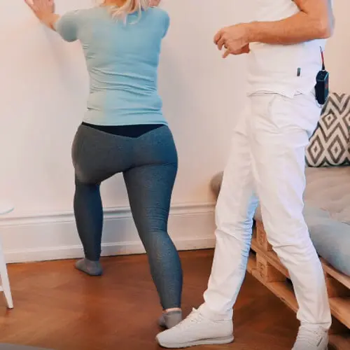 Eine Frau steht an einer Wand, stützt sich mit den Armen ab. Ein Bein steht leicht gebeugt an der Wand, das andere weit weggestreckt nach hinten. Roland Liebscher-Bracht steht hinter ihr. Die Übungen lindert Schmerzen bei einer Plantarfasziitis und Haglundferse.