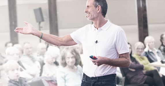 Auf diesem Bild sieht man Roland von Liebscher & Bracht . Er hält drei Stifte in der linken Hand mit den Farben Rot,Schwarz,Grün. Mit der rechten Hand zeigt er auf sitzende Personen die ihm zuhören. Roland ist ausgestattet mit einem Mikrofon und trägt ein weißes T-Shirt.