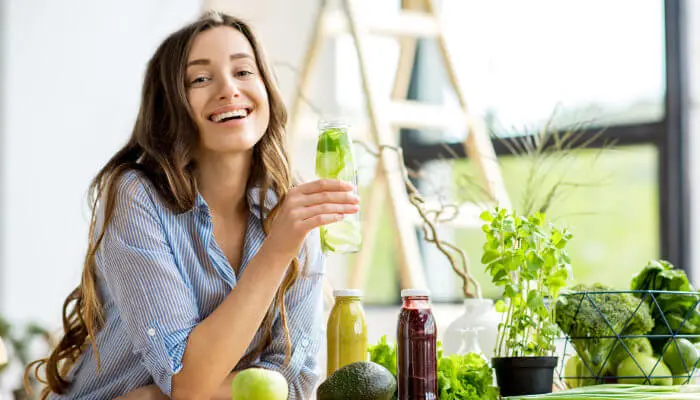 Eine lächelnde Frau trinkt einen grünen Smoothie und ist umringt von Gemüse