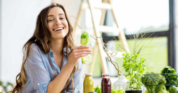 Eine lächelnde Frau trinkt einen grünen Smoothie und ist umringt von Gemüse