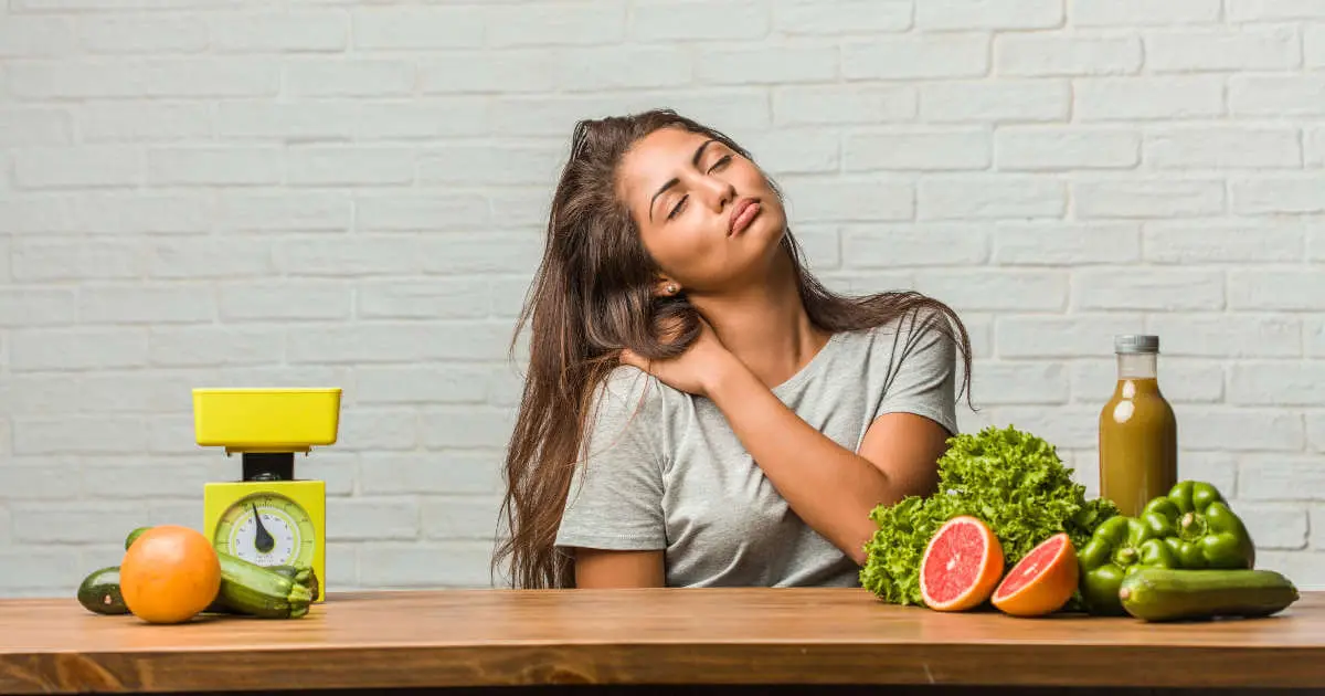 Eine Frau sitzt mit geschlossenen Augen an einem Tisch und berührt mit einer Hand ihre rechte Schulter. Auf dem Tisch liegen Paprika, Grapefruit, Zucchini, Orangen, Saft und eine Küchenwaage verteilt.
