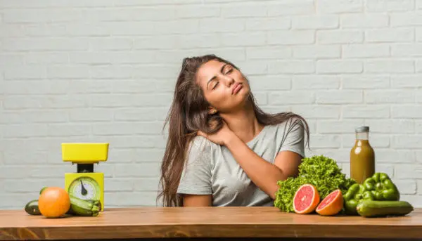 Eine Frau sitzt mit geschlossenen Augen an einem Tisch und berührt mit einer Hand ihre rechte Schulter. Auf dem Tisch liegen Paprika, Grapefruit, Zucchini, Orangen, Saft und eine Küchenwaage verteilt.