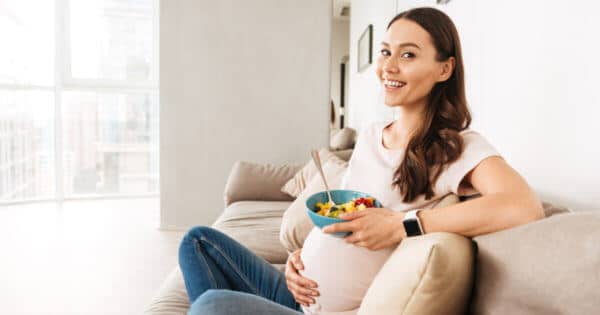 Eine schwangere, lächelnde Frau sitzt auf ihrem Sofa und hält eine Obstschale in der Hand