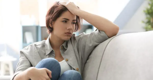 Eine Frau mit müdem Gesichtsausdruck sitzt mit angezogenen Knien auf einem Sofa. Ihre linke Hand hat sie auf dem Kopf abgelegt.
