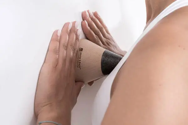 Zwei Frauenhände halten den "Drücker" mit der flachen Seite an die Wand und mit der Spitze drückt sie sich einen Punkt in Brusthöhe