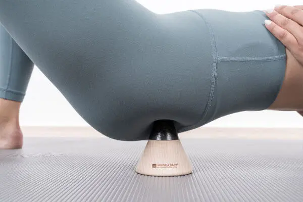 Eine Frau mit eng anliegender Hose sitzt auf dem kegelförmiges Sockelelement und drückt sich mit Spitze einen Punkt auf dem Po.