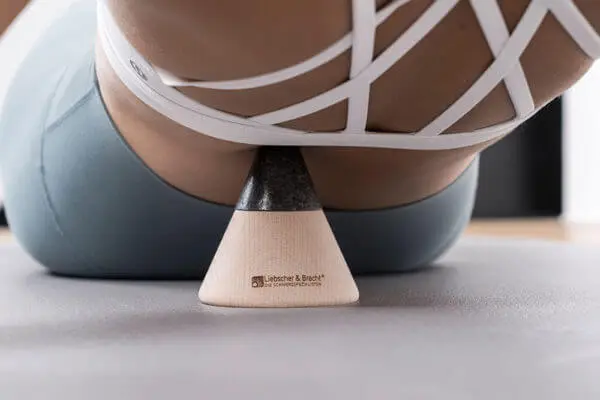 Eine Frau mit eng anliegender Hose liegt auf dem kegelförmiges Sockelelement und drückt sich mit der Spitze einen Punkt auf dem Rücken