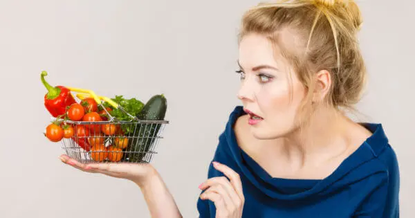 Eine Frau mit hochgesteckten Haaren blickt seitlich zu einem kleinen Einkaufskorb auf ihrer Handfläche. In diesem liegen Tomaten, Paprika, eine Gurke und grüne Blätter.