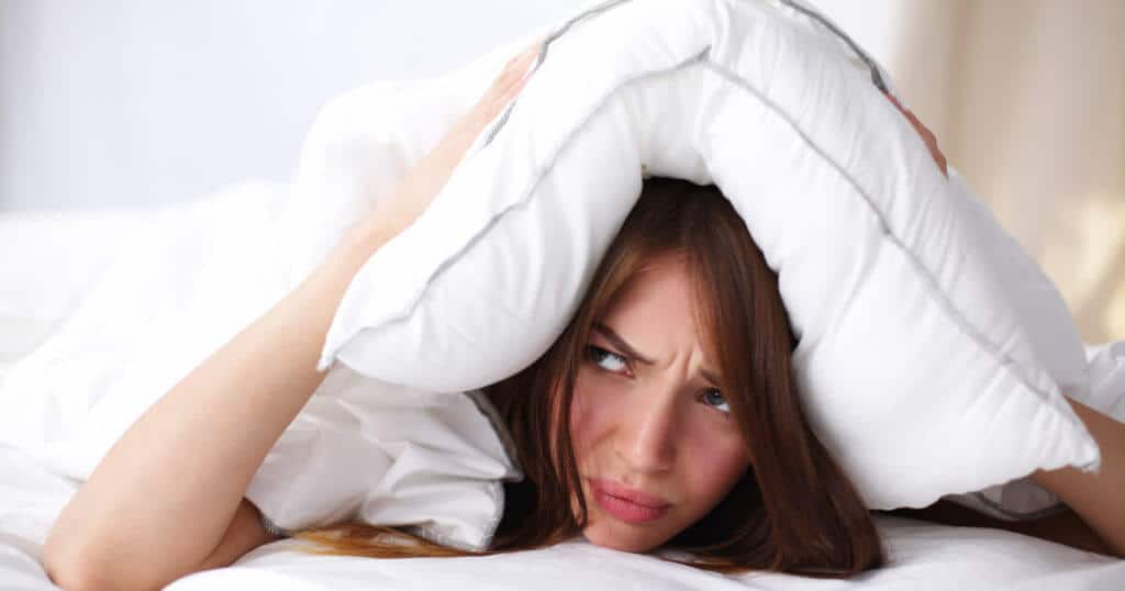 Eine Frau liegt auf einer Matratze und drückt ein Kissen über ihren Kopf. Dabei schaut sie etwas mürrisch zur Seite.