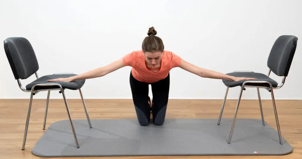 Eine Frau kniet auf einer Übungsmatte und stützt beide augestreckten Arme seitlich auf Stühlen ab, um das Zwerchfell zu dehnen.