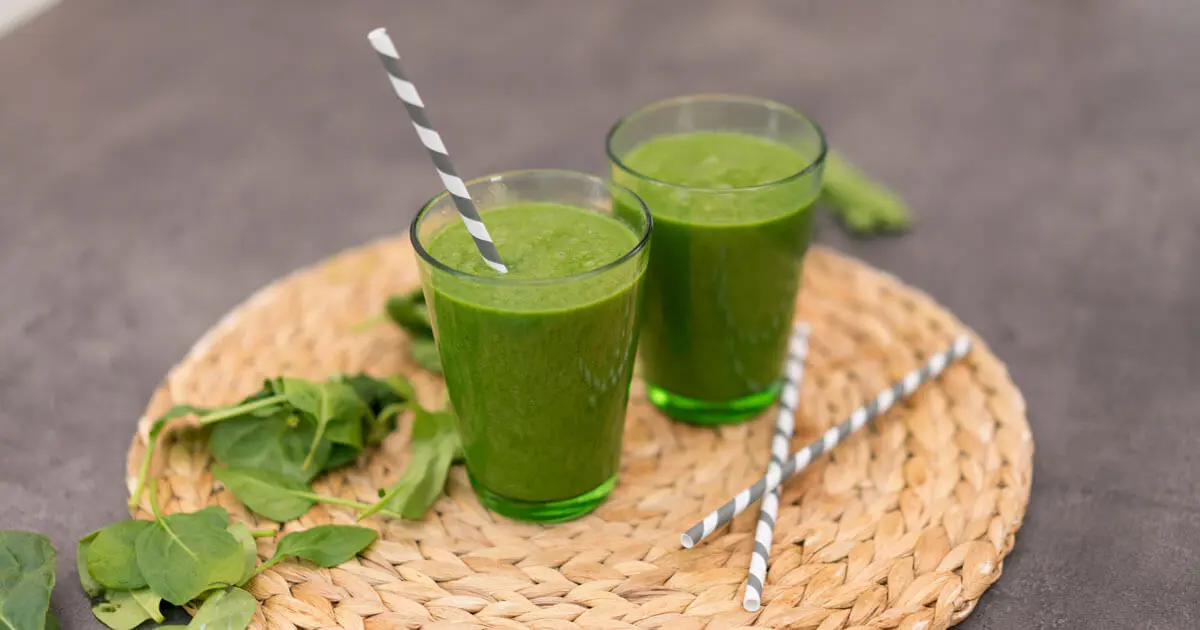 Zwei kleine Gläschen mit einem grünen Smoothie auf einem Untersetzer, dekoriert mit Kräutern und Strohhälmen