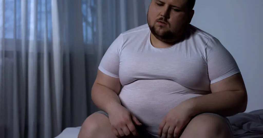 Ein dicker Mann sitzt unglücklich auf seinem Bett und fühlt sich müde und antriebslos