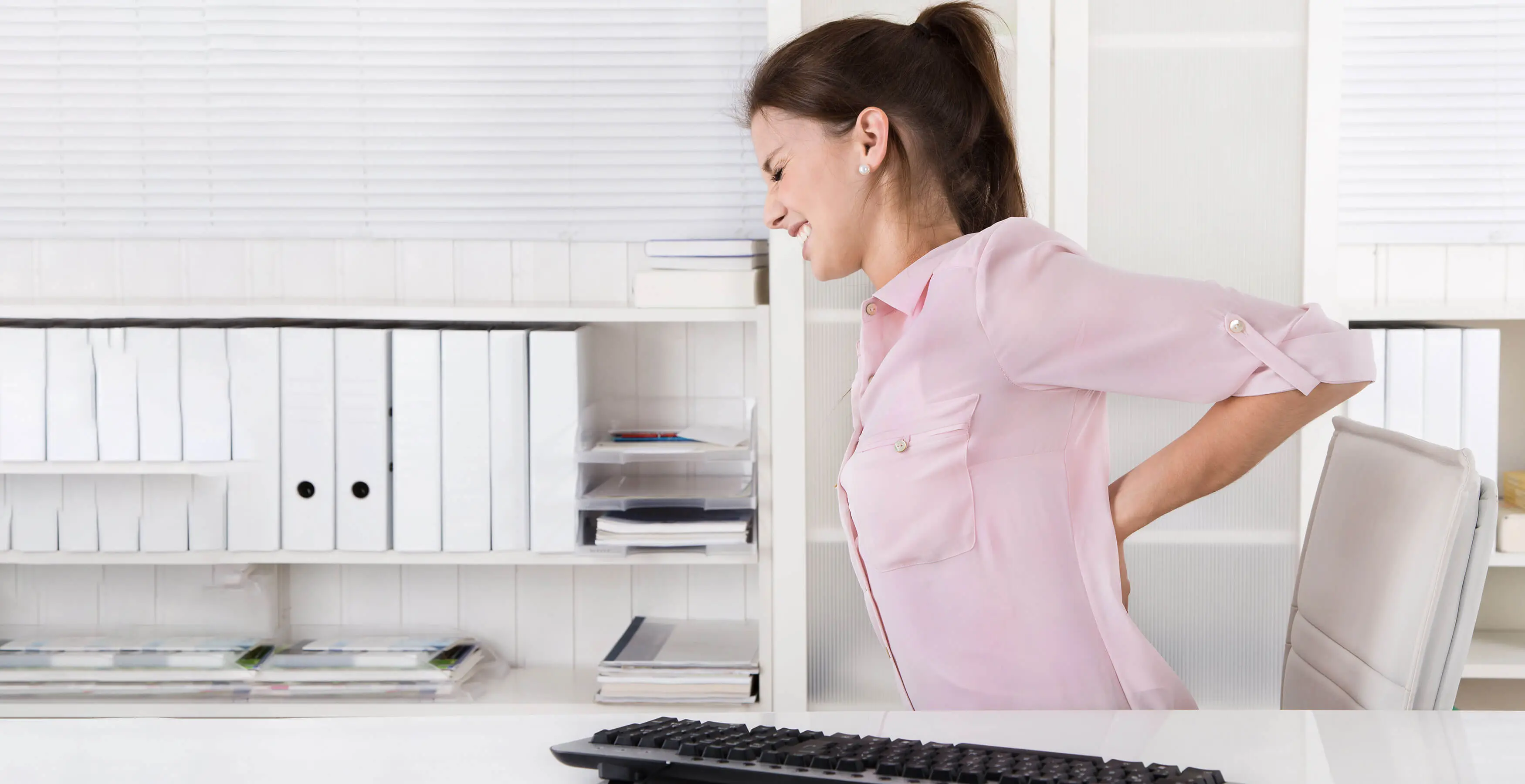 Eine Frau sitzt am Schreibtisch und stuetzt sich wegen ihrer Schmerzen den unteren Ruecken.