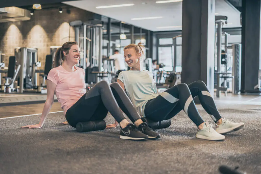 Zwei Frauen sitzen mit angewinkelten Beinen und nach hinten ausgestreckten Armen auf Medi-Faszienrollen von Liebscher & Bracht, die auf dem Boden liegen. Im Hintergrund sind Fitnessgeräte eines Studios zu sehen.