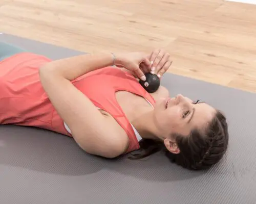 Eine Frau liegt auf einer Übungsmatte und führt eine Faszienrollmassage mit der Mini-Faszienkugel von Liebscher & Bracht durch. Mit beiden Händen drückt sie die Kugel von vorne in den Schulterbereich.