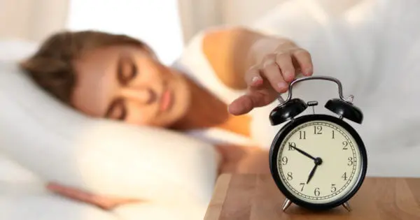 Eine Frau liegt im Bett, im Vordergrund steht ein klassischer Analogwecker. Sie tippt mit geschlossenen Augen auf ihn.