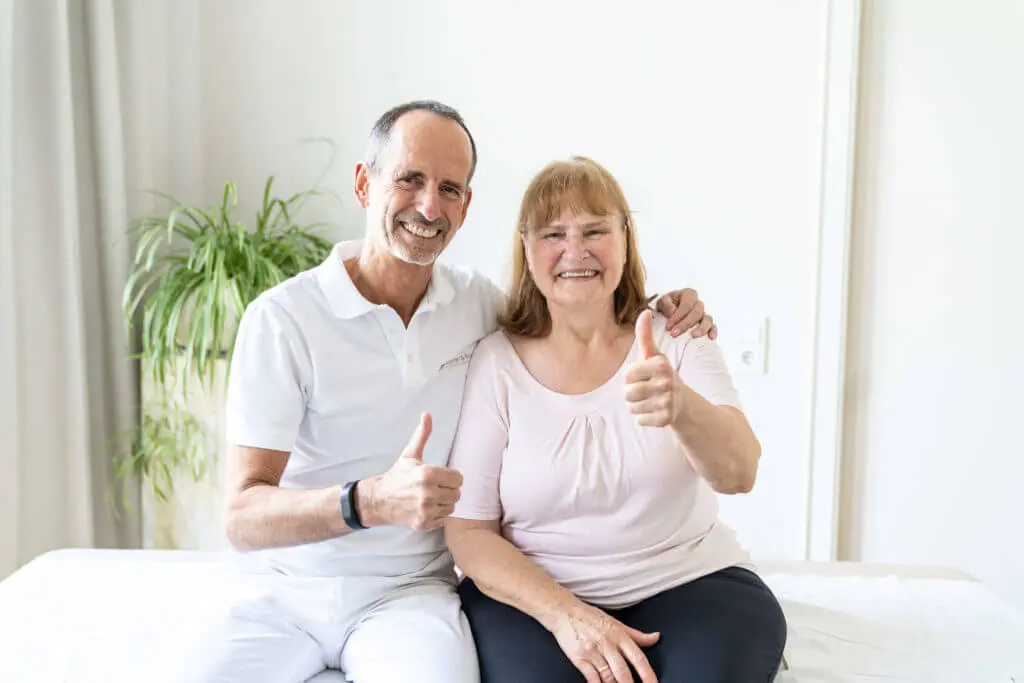 Roland Liebscher-Bracht und eine Patientin sitzen nach einer erfolgreichen Behandlung nebeneinander. Sie zeigen beide einen Daumen-hoch