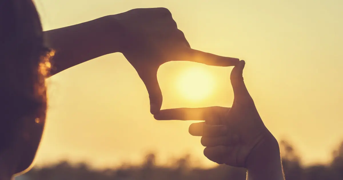 Zu sehen ist eine Frau, die beide Hände nutzt, um mit Daumen und Zeigefingern einen Rahmen zu formen und die Sonne "einzufangen".