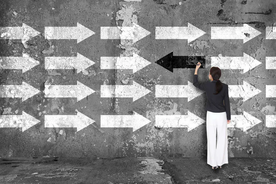 Eine Frau steht vor einer grauen Betonwand, auf der viele weiße Pfeile, die nach rechts zeigen, aufgemalt sind. Die Frau malt mit einem schwarzen Stift einen Pfeil, der entgegensetzt zu den anderen Pfeilen nach links zeigt.