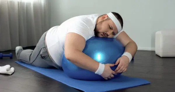 Ein uebergewichtiger Mann liegt auf einem Fitness-Ball und umklammert ihn, um sich noch halbwegs oben zu halten. Er hat einen lustlosen Ausdruck im Gesicht.