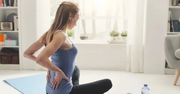 Eine Frau sitzt im Schneidersitz auf einer Matte und hälte ihre Hände an den unteren Rücken. Im Hintergrund sieht man ein freundliches Wohnzimmer.