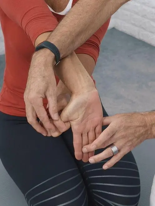 Eine Frau sitzt im Fersensitz auf dem Boden und streckt ihren rechten Arm aus, damit sie ihren Daumen der rechten Hand mit der linken Hand nach hinten ziehen kann, damit sie so ihre Daumenschmerzen loswerden kann