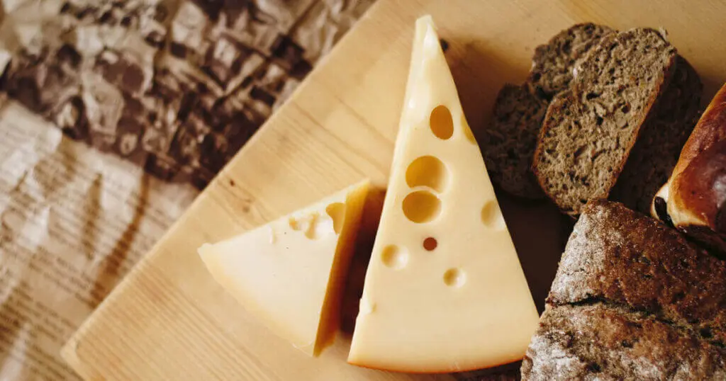 Zwei grobe Stücke Käse liegen gemeinsam mit etwas Brot auf einem Holzbrett.