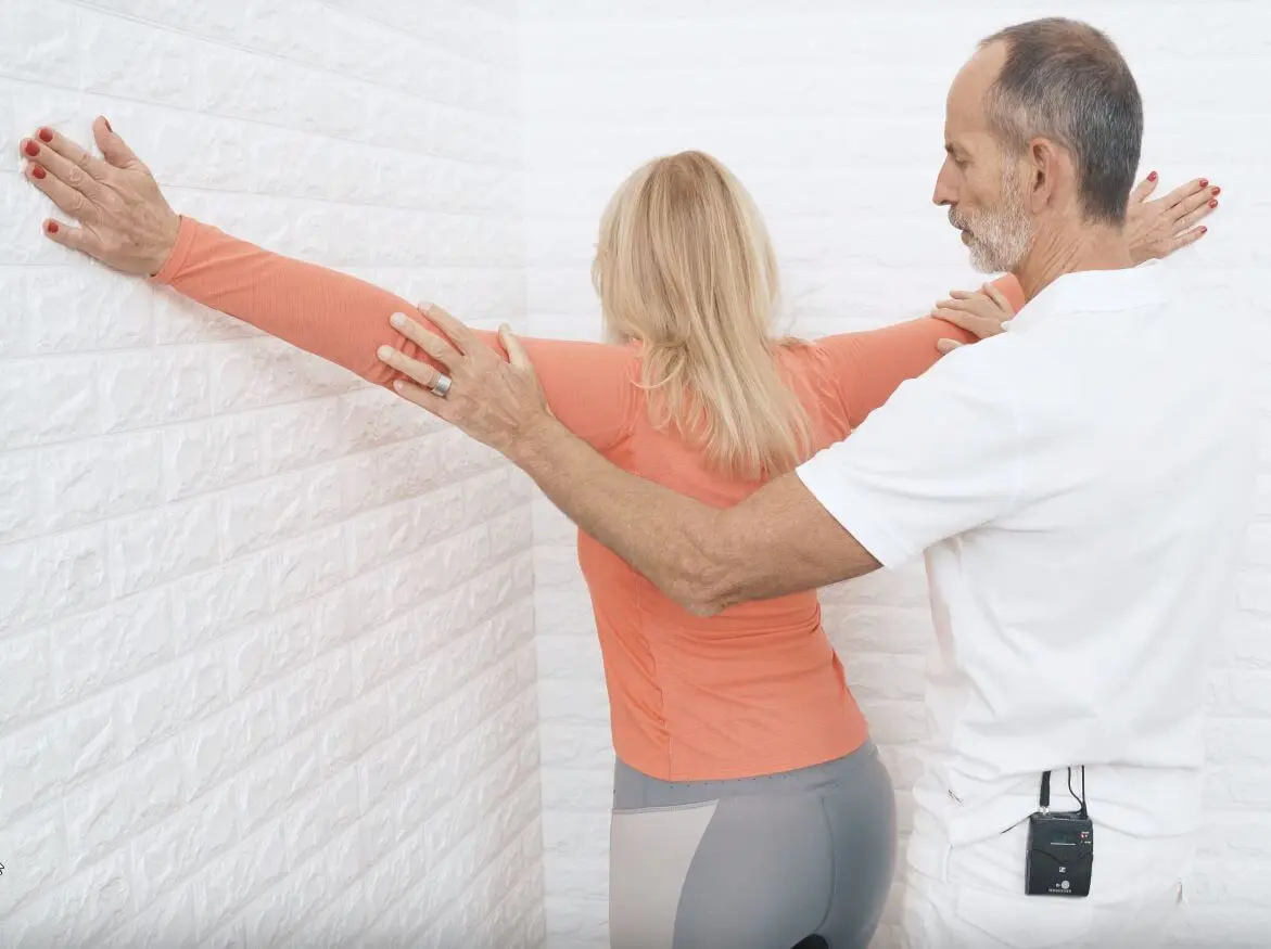 Eine Patientin steht mit erhobenen Armen in einer Zimmerecke und dehnt ihren Brustbereich auf. Ihre Arme sind etwas über Schulterhöhe voll ausgestreckt und Roland Liebscher-Bracht leitet sie an.