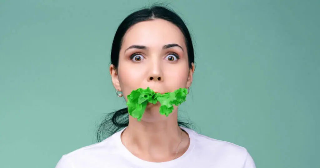 Eine junge Frau hat ein grünes Salatblatt im Mund und schaut erschrocken in die Kamera