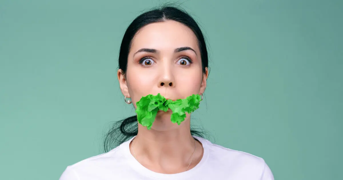 Eine junge Frau hat ein grünes Salatblatt im Mund und schaut erschrocken in die Kamera