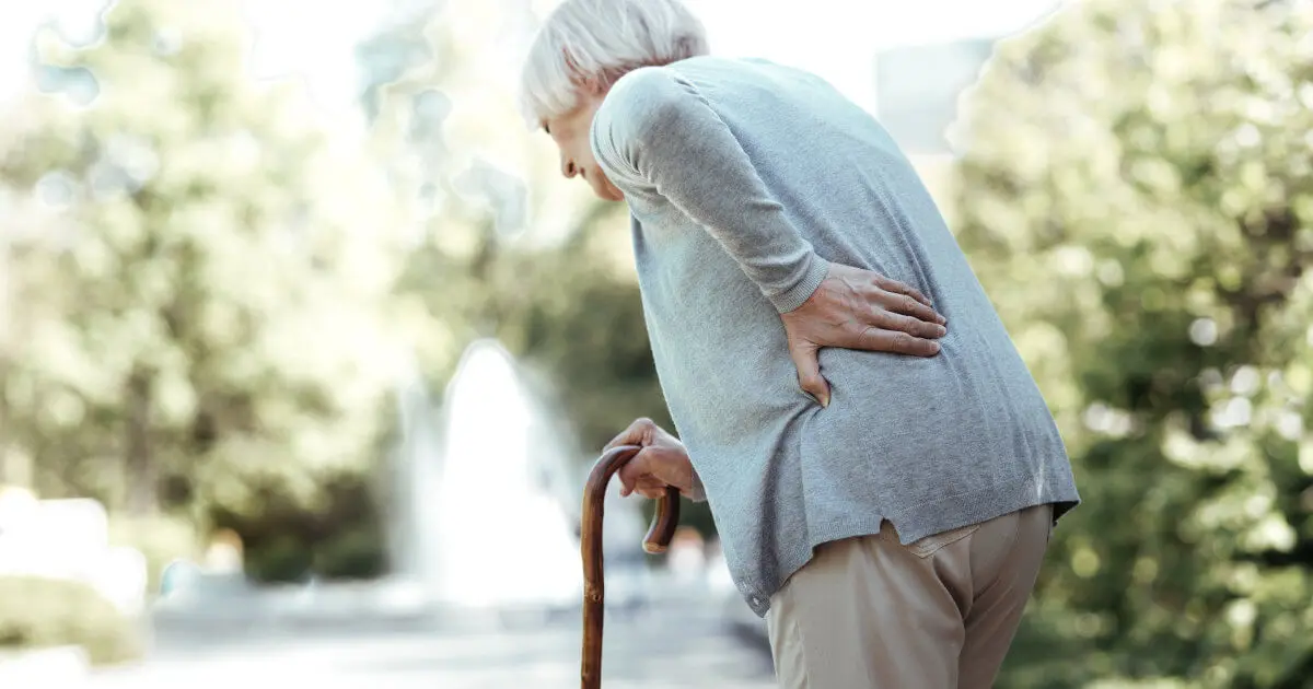 Eine aeltere Frau stuetzt sich auf ihren Gehstock. Sie kruemmt sich vor Schmerzen und fasst sich mit ihrer linken Hand an den unteren Ruecken.
