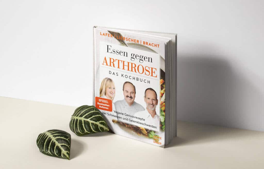 Das Kochbuch mit Schmerzspezialist Roland Liebscher-Bracht, Dr. med. Petra Bracht sowie Starkoch Johann Lafer auf dem Cover ist aufgestellt. Vor dem Buch liegen zwei grüne große Blätter.