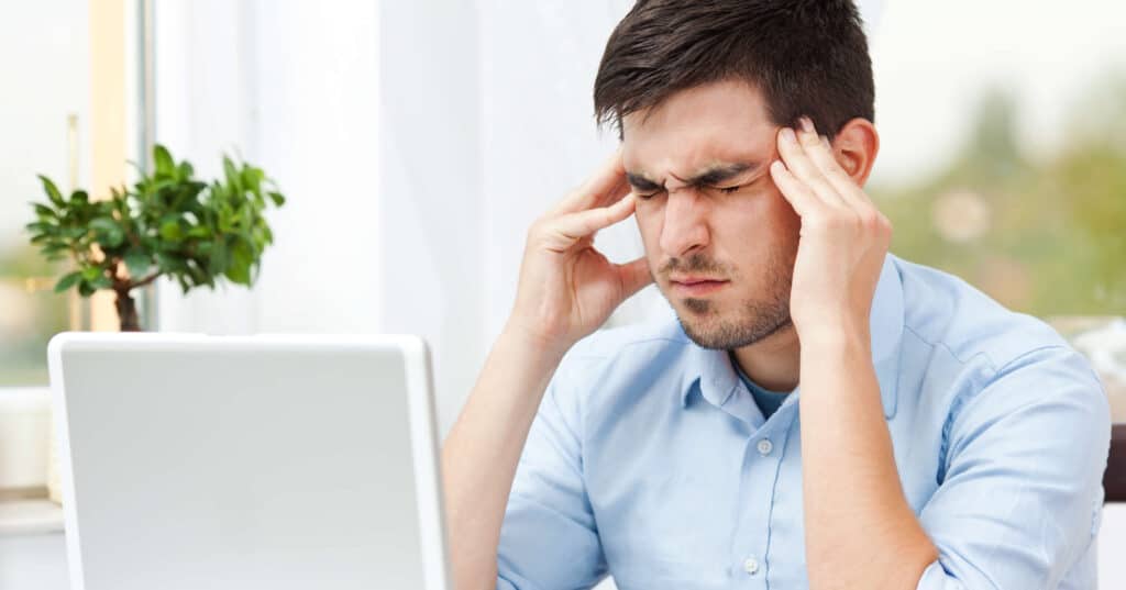 Ein Mann sitzt mit geschlossenen und verkrampftem Gesicht da, dabei berührt er seine Schläfen beidseitig mit den Händen.