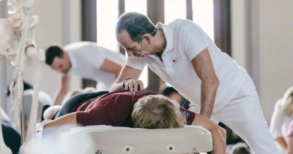 Schmerzspezialist Roland Liebscher-Bracht behandelt im Rahmen der Schmerztherapie-Ausbildung einen Teilnehmer per Osteopressur. Der Teilnehmer befindet sich auf einer Liege, Roland Liebscher-Bracht steht rechts davon und drückt mithilfe seines Ellenbogens einen Osteopressurpunkt im unteren Rücken des Teilnehmers.