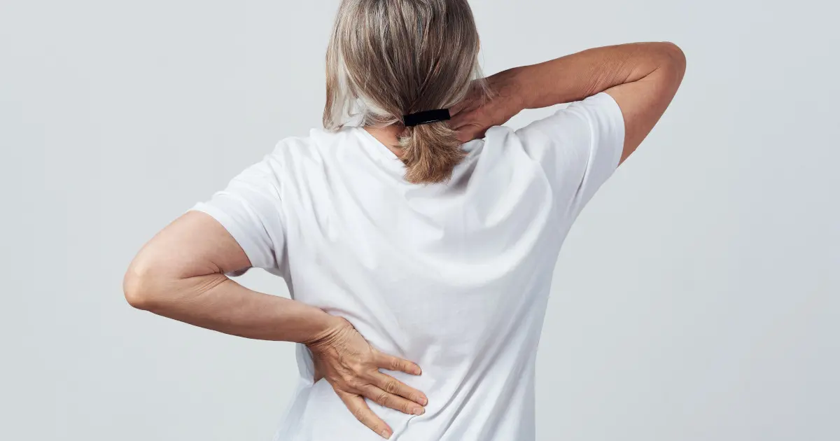 Zu sehen ist eine Frau von hinten. Mit ihrer linken Hand hält sie sich den Rücken, mit der rechten den Nacken.