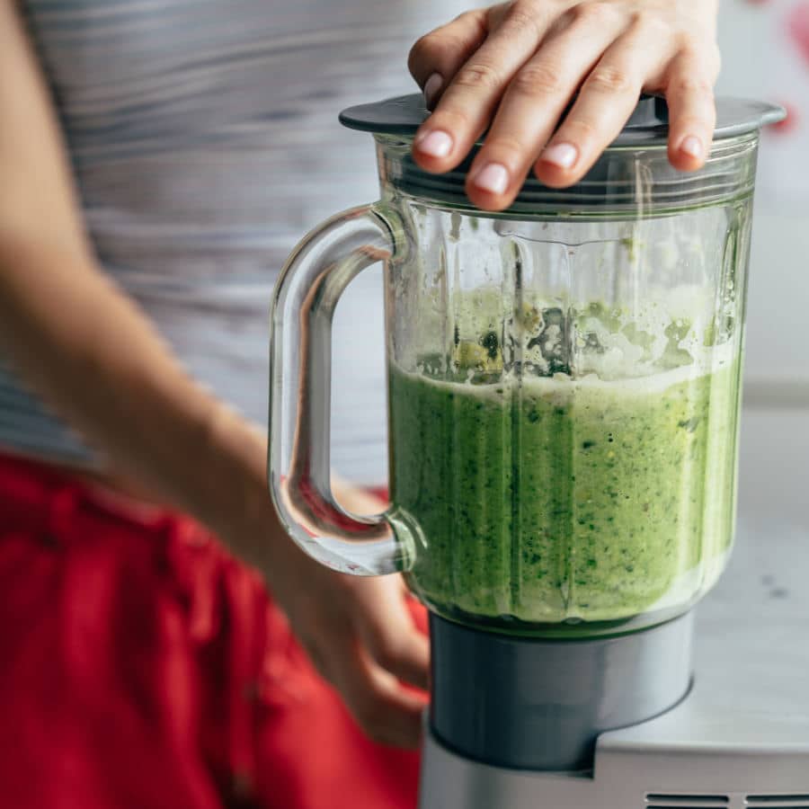 Eine junge Frau mischt sich in einem Mixer einen grünen Smoothie zusammen. Der Mixer steht im Fokus