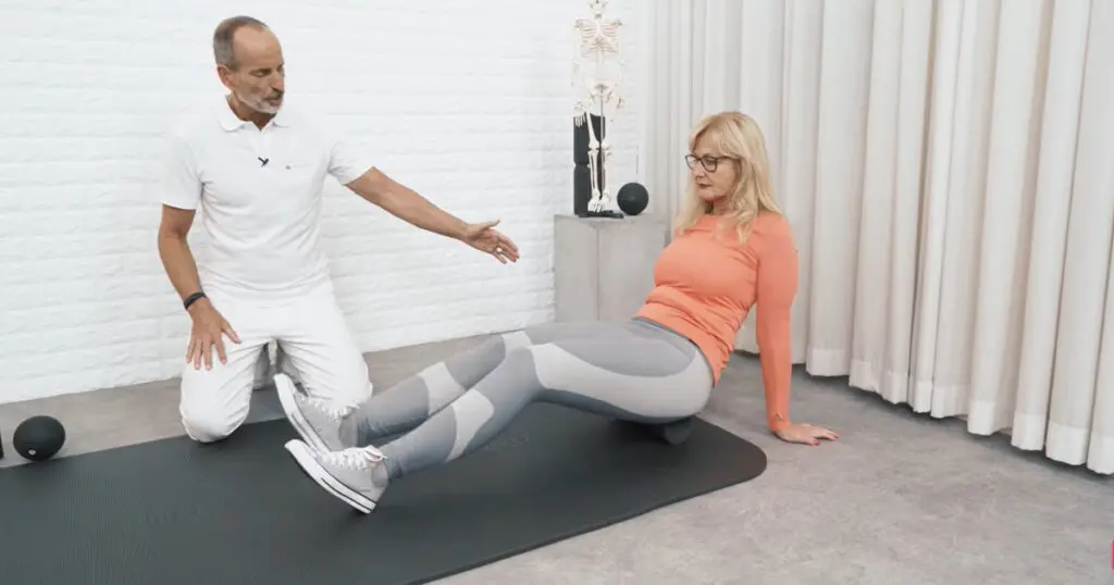 Eine Frau sitzt mit dem Steißbein auf einer Faszienrolle, um eine Faszien-Rollmassage für den gesamten Rücken durchzuführen