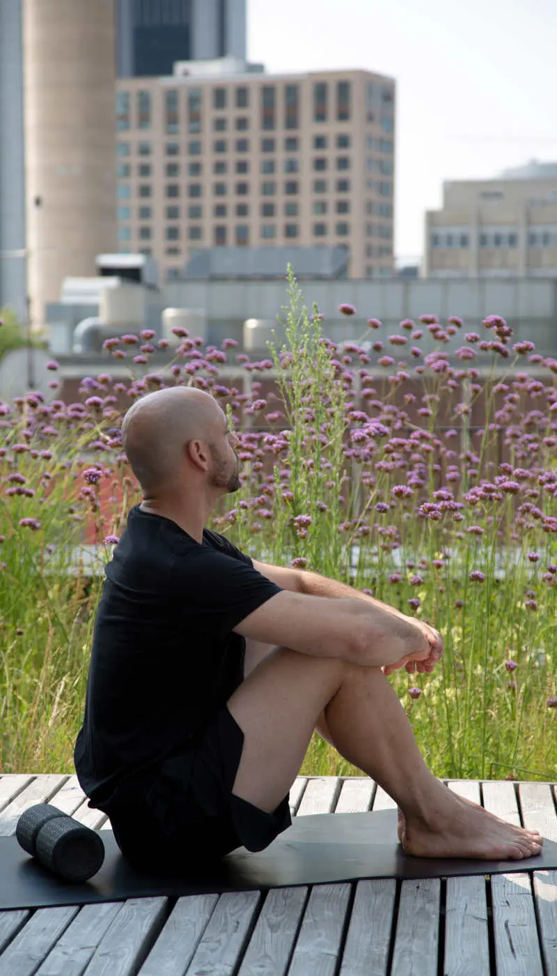 Zu sehen ist ein Mann, der auf einer Holzterrasse vor einer Blumenwiese mit Blick auf die Stadt sitzt. Hinter ihm befindet sich eine Faszienrolle.