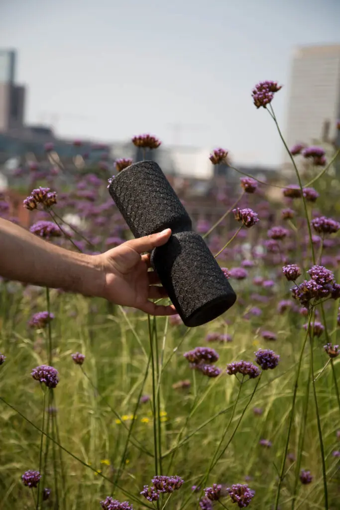 Zu sehen ist eine Hand, die eine Faszienrolle mit Wirbelsäulenvertiefung in die Kamera hält. Im Hintergrund ist eine Blumenwiese mit lila Blumen zu sehen.