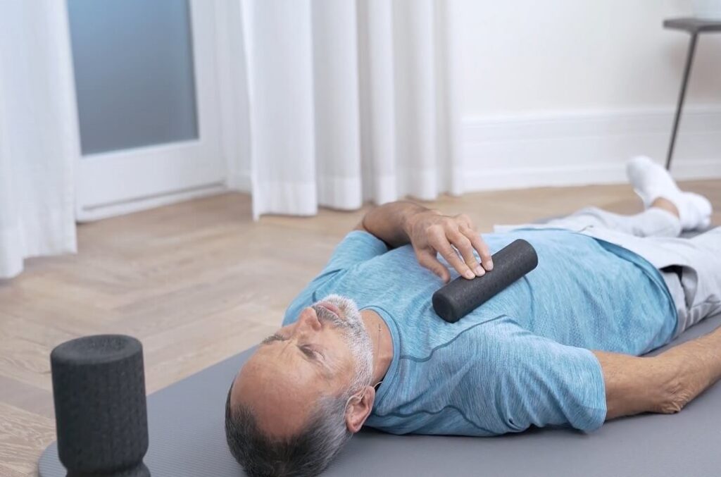 Ein Mann liegt auf einer Übungsmatte und rollt einhändig mit einer Faszienrolle über seinen Brustmuskel