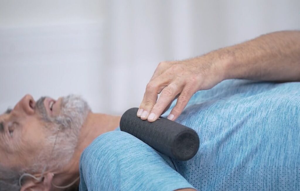 Nahaufnahme eines Mannes, der eine Faszienrollmassage im Brustbereich durchführt und die Faszienrolle in der rechten Achselhöhle einhändig hält.
