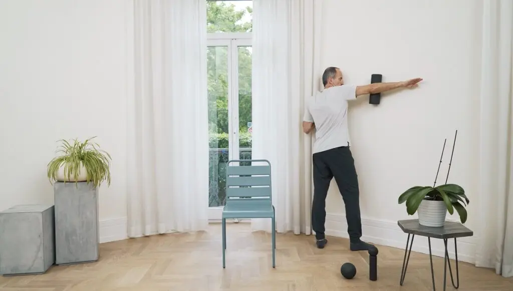 Ein Mann steht in einem Raum zwischen Tisch und Stuhl. Er drückt seinen Arm an der Wand auf eine Faszienrolle, um eine Faszien-Rollmassage für die Schultern durchzuführen