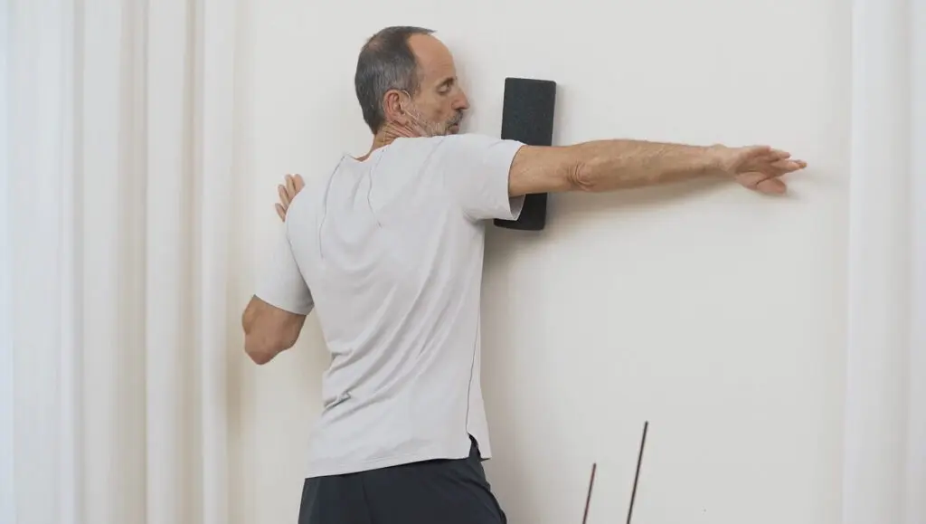 Ein Mann steht in einem Raum und drückt seinen Arm an der Wand auf eine Faszienrolle und rollt auf diese Weise in Richtung Schulter
