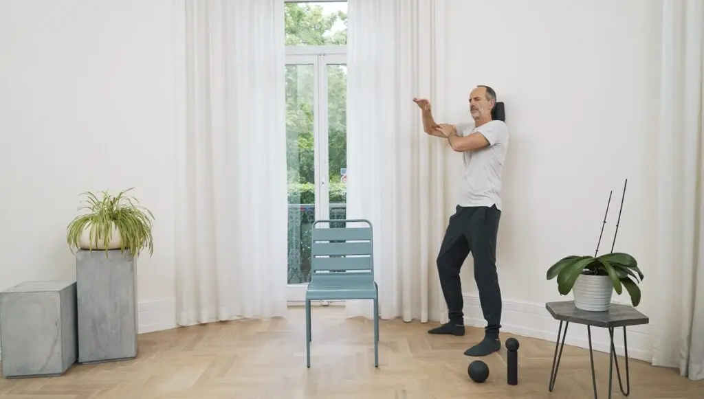 Ein Mann steht in einem Raum zwischen einem blauen Stuhl und einem Tisch und drückt mit seiner Schulter eine Faszienrolle an die Wand